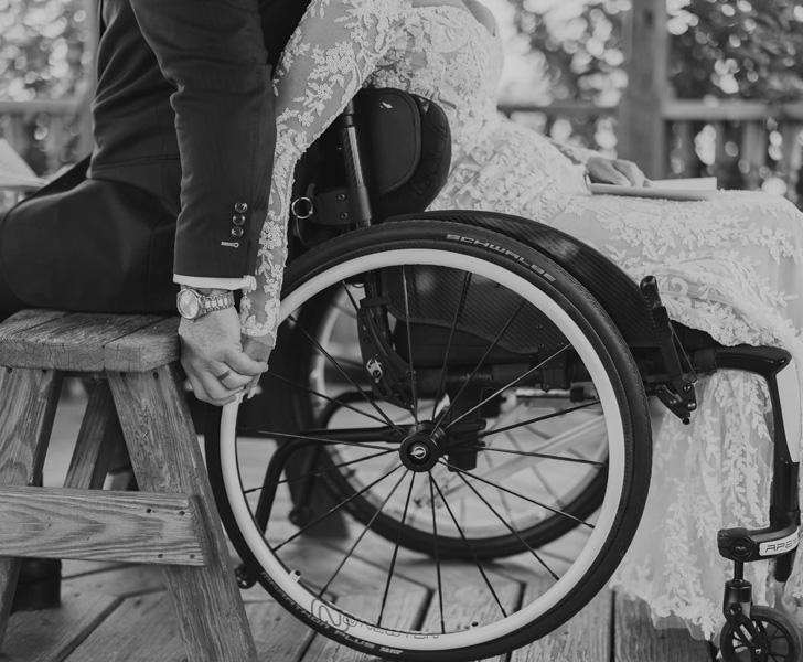 Couple sitting back-to-back wearing their wedding attires. The bride is using a wheelchair.
